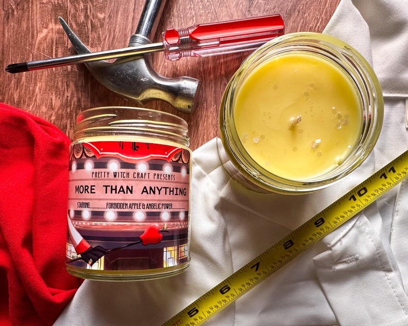 May include: A glass jar candle with yellow wax and a label that reads "PRETTY WITCH CRAFT PRESENTS MORE THAN ANYTHING STARRING FORBIDDEN APPLE & ANSELIC POWER".