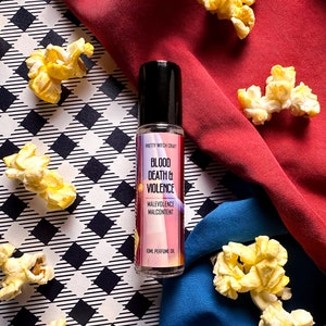 May include: A small glass bottle of perfume oil with a black cap. The bottle is labelled "Pretty Witch Craft" and "Blood Death & Violence" with the subtext "Malevolence Malcontent". The bottle is sitting on a black and white chequered surface with popcorn scattered around it.