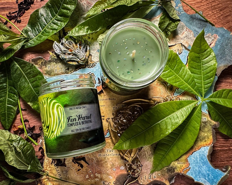 May include: Two green soy blend candles with a map and green leaves in the background. The candle on the left has a label that reads "Fen Harel Temples & Betrayal".