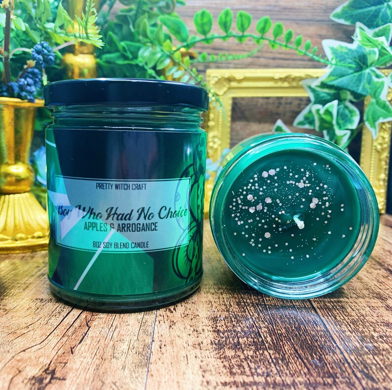 May include: A green soy blend candle with a black label that reads "Sor The Had No Choice Apples & Arrogance 8oz Soy Blend Candle". The candle is in a clear glass jar with a black lid. The candle is sitting on a wooden surface.