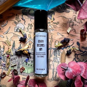 May include: A small, clear glass bottle with a black roll-on top. The bottle is labelled "Pretty Witch Craft" and contains a perfume oil called "Death Becomes Him: Necropolis Garden Old Tomes". The bottle is sitting on a brown surface with a variety of dried flowers and other botanicals.