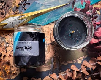 May include: A black soy blend candle in a glass jar with a label that reads "Dread Wolf Tears & Lies". The candle is surrounded by dried leaves and a gold and blue decorative object.