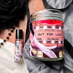 May include: A glass candle jar with a red and white label that reads "Pretty Witch Craft Presents Out For Love Starring Protection & Regret". The candle jar is sitting on a white surface with a black and white patterned fabric.