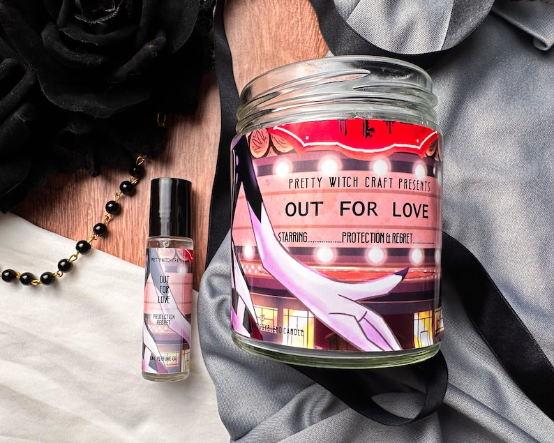 May include: A glass candle jar with a red and white label that reads "Pretty Witch Craft Presents Out For Love Starring Protection & Regret". The candle jar is sitting on a white surface with a black and white patterned fabric.