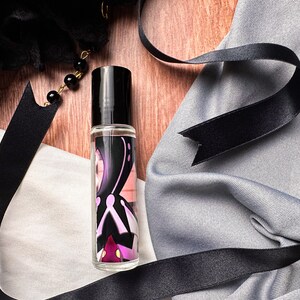 May include: A clear glass roller bottle with a black cap and a colorful design featuring a purple, pink, and white abstract pattern. The bottle is sitting on a wooden surface with a black ribbon and fabric.
