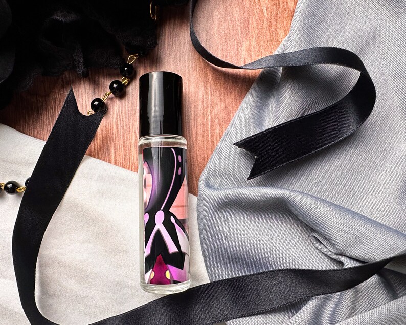 May include: A clear glass roller bottle with a black cap and a colorful design featuring a purple, pink, and white abstract pattern. The bottle is sitting on a wooden surface with a black ribbon and fabric.