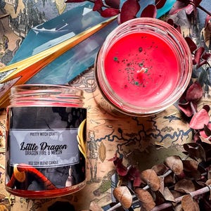May include: Two glass jar candles with red wax, one open and one closed, are on a map-themed surface. The label on the closed candle reads "Little Dragon Dragon Fire & Melon 8oz Soy Blend Candle". Dried leaves and a fantasy illustration complete the scene.