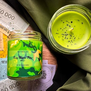 Puede incluir: Un tarro de vela de vidrio verde con una etiqueta verde y amarilla que dice "Pretty Witch Craft Buy Yours Now Money 8oz Soy Blend Candle". La vela está rodeada de billetes de dinero falsos.