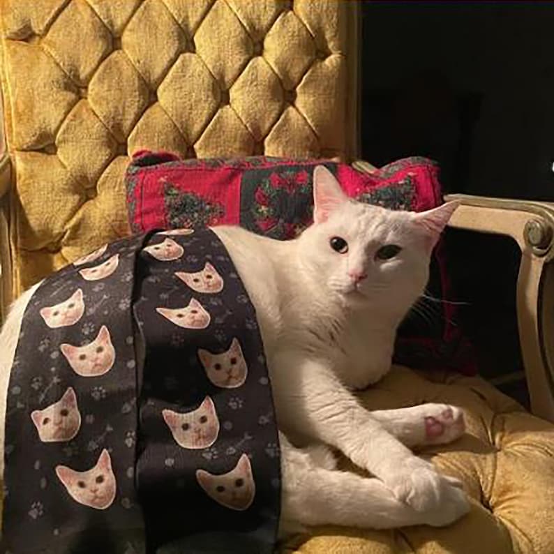 May include: A white cat with pink paw pads resting on a yellow tufted chair. The cat is partially covered by a black fabric strip featuring a repeating pattern of white cat faces and small gray paw prints, set against a red patterned pillow.