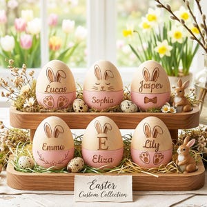 May include: Wooden Easter egg ornaments with bunny ears and personalized names. Each egg has a pink base and is displayed on a wooden tray with decorative grass and small bunny figurines. The text "Easter Custom Collection" is also visible.