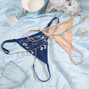 May include: Two delicate lace lingerie sets with a silver chain and charm necklace featuring the word "AQUARIUS". One set is navy blue and the other is beige.