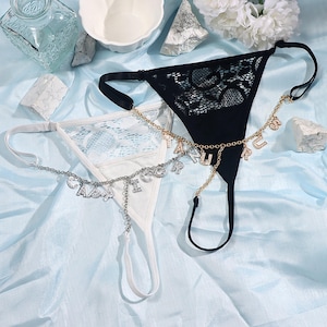 May include: Two black and white lace lingerie sets with gold and silver chains with letter charms. The chains are attached to the lingerie and have charms that spell out "TAN" and "CADDY".
