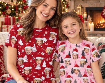 May include: Two short-sleeved t-shirts featuring a repeating pattern of faces wearing Santa hats, Christmas trees, and snowflakes. The left shirt is red with white graphics, and the right shirt is light pink with dark pink and white graphics.