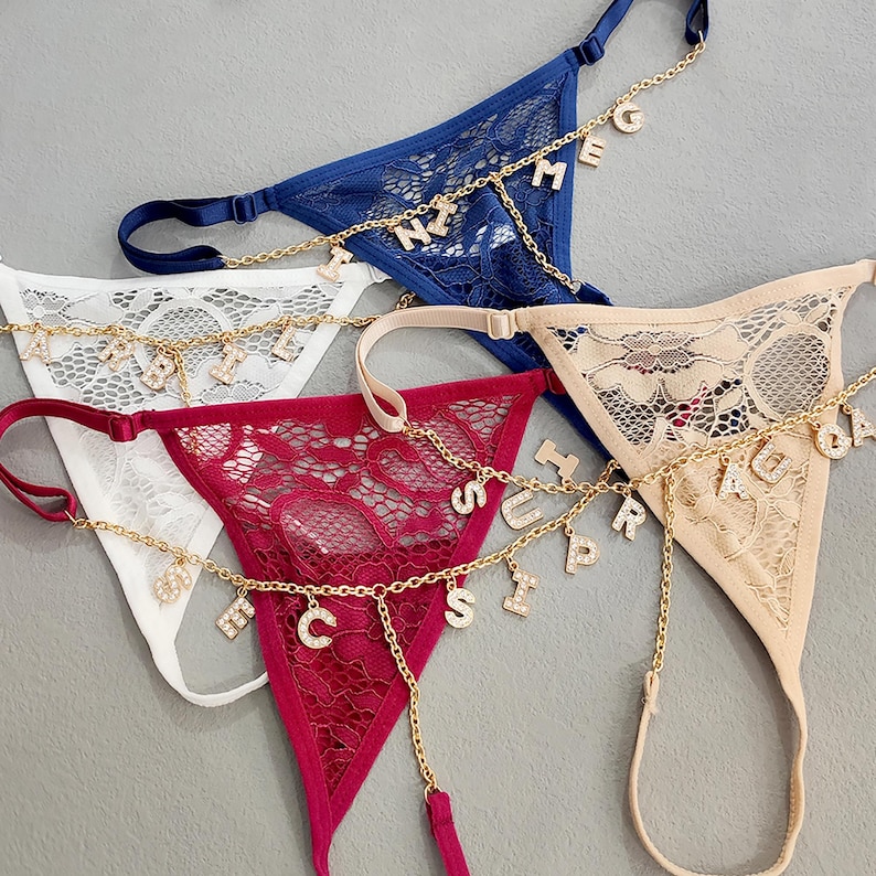 May include: Three pairs of lacy underwear with gold chains and letter charms. The chains are attached to the underwear and have charms with letters spelling out words. The underwear is in white, red, and beige.
