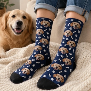 May include: A pair of navy blue crew socks featuring a repeating pattern of golden retriever faces, white paw prints, and white bone shapes. The socks have solid black toe and heel sections and are worn on the lower legs with blue denim jeans.