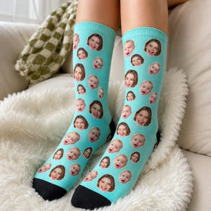 May include: A pair of light blue crew socks featuring a repeating pattern of various photographic portraits of a baby and a young adult. The socks have a solid black toe section and a solid black heel section, providing a distinct contrast to the blue.