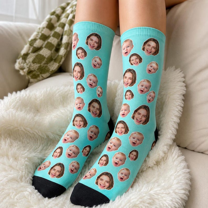 May include: A pair of light blue crew socks featuring a repeating pattern of various photographic portraits of a baby and a young adult. The socks have a solid black toe section and a solid black heel section, providing a distinct contrast to the blue.