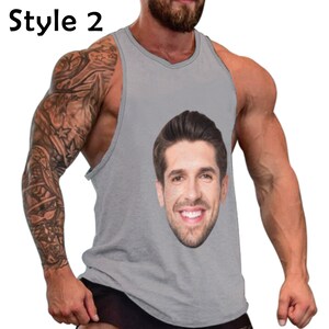May include: A gray sleeveless tank top with a man's face printed on the front. The face is smiling and has brown hair. The tank top is being worn by a person with tattoos on their arm.