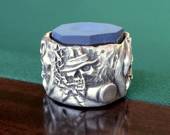 Puede incluir: Un portagises de taco de billar de color plateado con un diseño detallado de calavera. El soporte contiene una pieza de tiza azul octogonal. El fondo muestra una mesa de billar verde y varias bolas de billar.