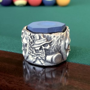 May include: A silver-coloured pool cue chalk holder featuring a detailed skull design. The holder contains a blue, octagonal piece of chalk. The background shows a green pool table and several billiard balls.