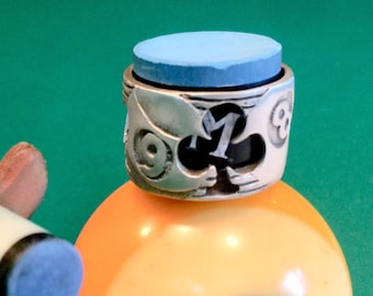 Könnte beinhalten: Eine gelbe Billardkugel mit der Nummer 9 darauf. Die Kugel liegt auf einem silbernen Ring mit einem blauen Filzpolster obenauf. Der Ring trägt die Nummer 9 und ein Kleeblatt.