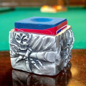 May include: A silver, skull-shaped,  pool cue chalk holder with a blue and red chalk cube on top.