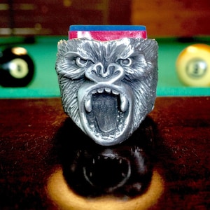 May include: A silver gorilla head pool cue rest with an open mouth and sharp teeth. The rest is sitting on a green felt pool table with two billiard balls in the background.