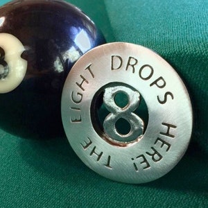 May include: A black eight ball with a white number eight on it is next to a silver metal disc with the words "The Eight Drops Here" engraved on it.