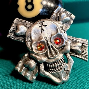 May include: A pewter skull with red and orange gem eyes, crossed over with two pieces of wood. The skull is detailed with a cracked surface and a toothy grin.