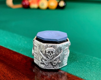 Puede incluir: Un porta tiza de billar de plata con un diseño de calavera y huesos cruzados. El porta tiza tiene un inserto de tiza azul.
