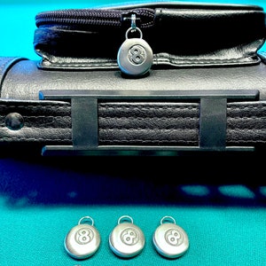 May include: Black leather pool cue case with a zipper closure and a silver 8-ball charm. The case has a black strap with a silver buckle. Three additional silver 8-ball charms are shown on a green felt surface.