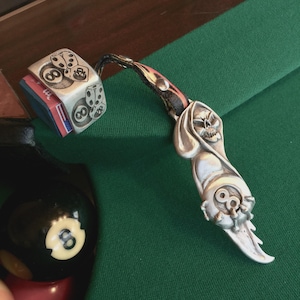 May include: A silver pool cue holder featuring a skull design and the number eight. It's connected to a dice-shaped chalk holder, also with the number eight and dice symbols. The items are displayed on a green felt pool table.
