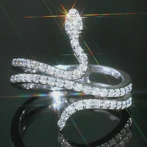 Peut inclure: Bague serpent argentée ornée de pierres précieuses claires et étincelantes. La tête du serpent est relevée et le corps s'enroule autour du doigt. La bague a une finition brillante, reflétant la lumière sur un fond sombre.