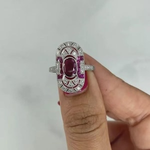 May include: An Art Deco-style ring featuring a large oval ruby center stone, surrounded by smaller rubies and clear accent stones. The ring has a silver-colored band and a detailed, geometric design. The ring's design is symmetrical and elegant.