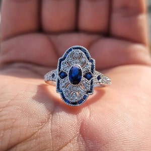 May include: A silver ring with a blue sapphire center stone surrounded by smaller diamonds. The ring has an intricate design with a filigree pattern.