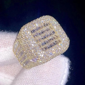 Könnte beinhalten: Ein goldener Ring mit einem großen quadratischen Mittelstück, das von kleineren Steinen umgeben ist. Das Mittelstück besteht aus Baguettesteinen.