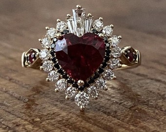 Könnte beinhalten: Ein goldener Ring mit einem großen roten herzförmigen Edelstein, umgeben von einem Heiligenschein aus kleineren weißen und schwarzen Edelsteinen.