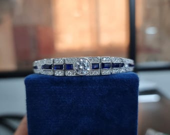 May include: A silver bracelet with a large round clear stone surrounded by smaller clear stones and blue stones. The bracelet is on a blue velvet box.