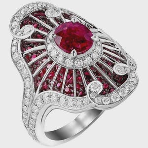 May include: An ornate, silver-toned ring featuring a large, round, ruby-red gemstone at the center, surrounded by smaller rubies and sparkling white diamonds. The ring has an Art Deco design with intricate detailing.