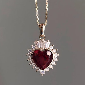 Può includere: Una collana con catena dorata con un ciondolo a forma di cuore rosso circondato da pietre di zircone cubico trasparenti.
