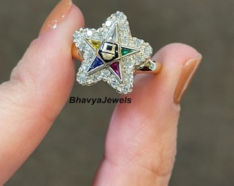 May include: A gold ring with a five-pointed star design. The star is set with small diamonds and has a smaller five-pointed star in the center with colored stones. The ring is on a finger.