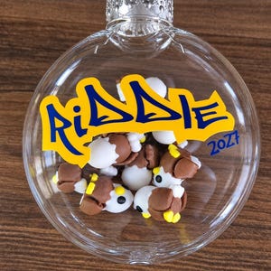 May include: A clear, spherical glass ornament filled with small, brown and white clay eagle figurines. The front of the ornament features the name "RIDDIE" printed in blue letters on a yellow background, with the year "2027" in blue text.