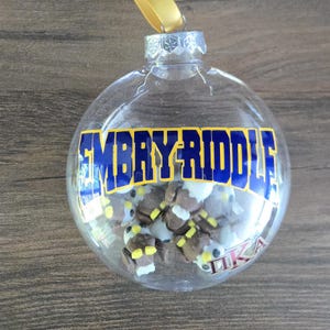 May include: Clear, round Christmas ornament with the text "EMBRY-RIDDLE" in blue and yellow. The ornament contains small, brown and white figures. A gold ribbon is attached for hanging. The letters "ΠKA" are visible on the ornament.