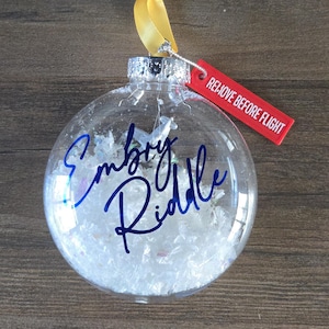 May include: Clear Christmas ornament with the name "Embry Riddle" in blue script. The ornament is filled with white, iridescent confetti and has a silver cap with a yellow ribbon. A red tag reads "REMOVE BEFORE FLIGHT."