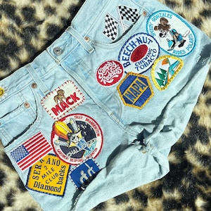 May include: Light blue denim shorts adorned with various embroidered patches. Patches include racing flags, a cartoon character, and brand logos. The shorts have a distressed look with a frayed edge.