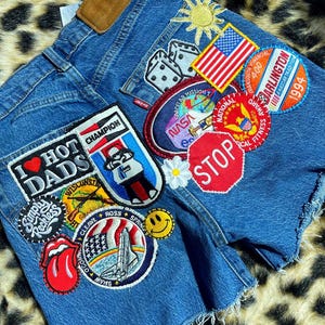 May include: Blue denim shorts with various embroidered patches, including a sun, dice, an American flag, and the text "I Hot Dads". The shorts have a frayed hem and a brown leather patch.