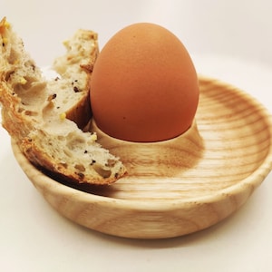 Peut inclure: Un œuf brun dans un porte-œuf en bois avec un morceau de pain à côté.