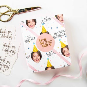 May include: A birthday gift wrapped in white paper with repeating images of a woman's face wearing a party hat. The wrapping paper has pink diagonal stripes, blue stars, and the words "22 Today" and "Happy Birthday". A pink sticker reads "You've Got Great Taste".