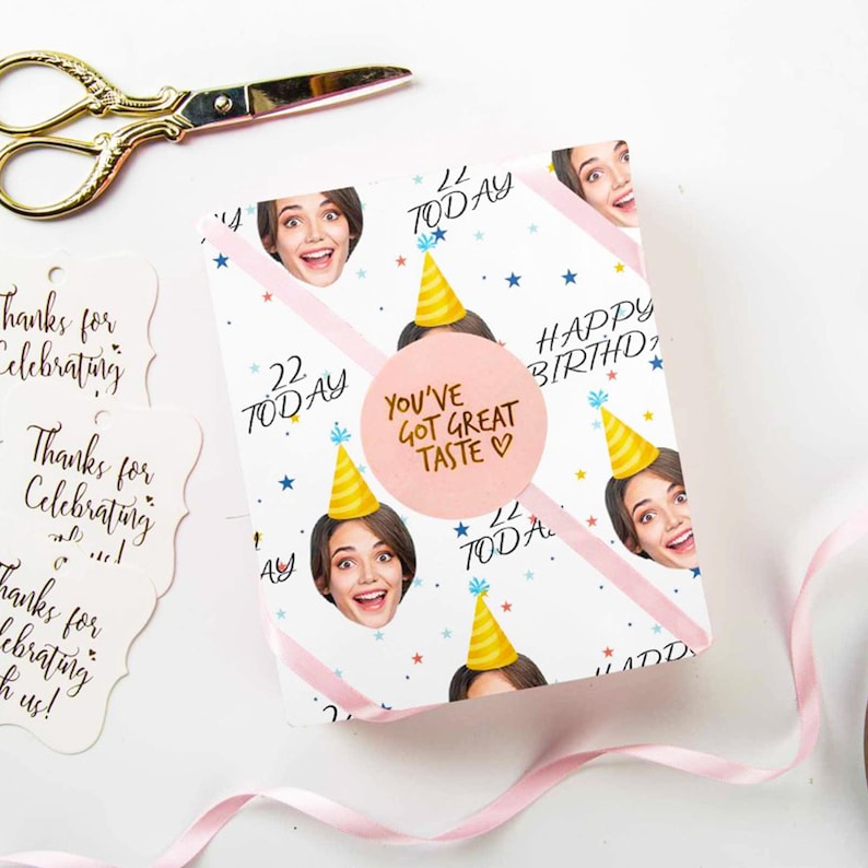 May include: A birthday gift wrapped in white paper with repeating images of a woman's face wearing a party hat. The wrapping paper has pink diagonal stripes, blue stars, and the words "22 Today" and "Happy Birthday". A pink sticker reads "You've Got Great Taste".
