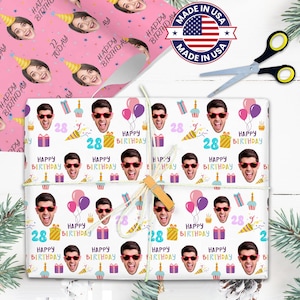 May include: Birthday gift wrap with a repeating pattern of a person's face wearing sunglasses, balloons, gifts, and the words "Happy Birthday" and the number "28". A roll of wrapping paper with a face wearing a party hat is visible. Scissors and a "Made in USA" sticker are also present.